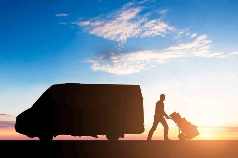 Silhouette Of Delivery Courier With Cardboard Boxes On Trolley Near The Van At Sunset Silhouette Of Delivery Courier With Cardboard Boxes On Trolley Near The Van At Sunset